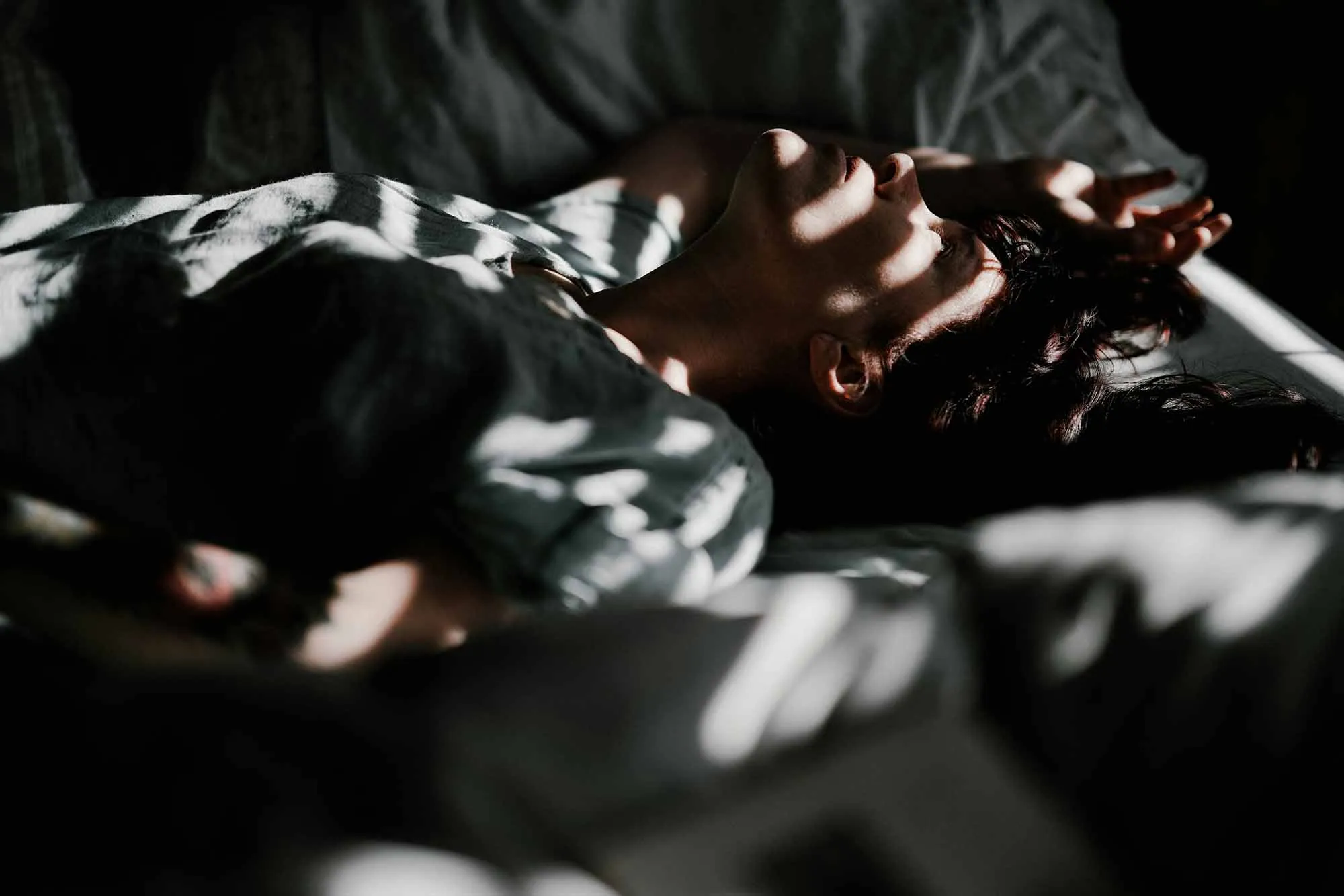 A woman lying in bed with soft shadows across her face, appearing restless and unable to sleep.