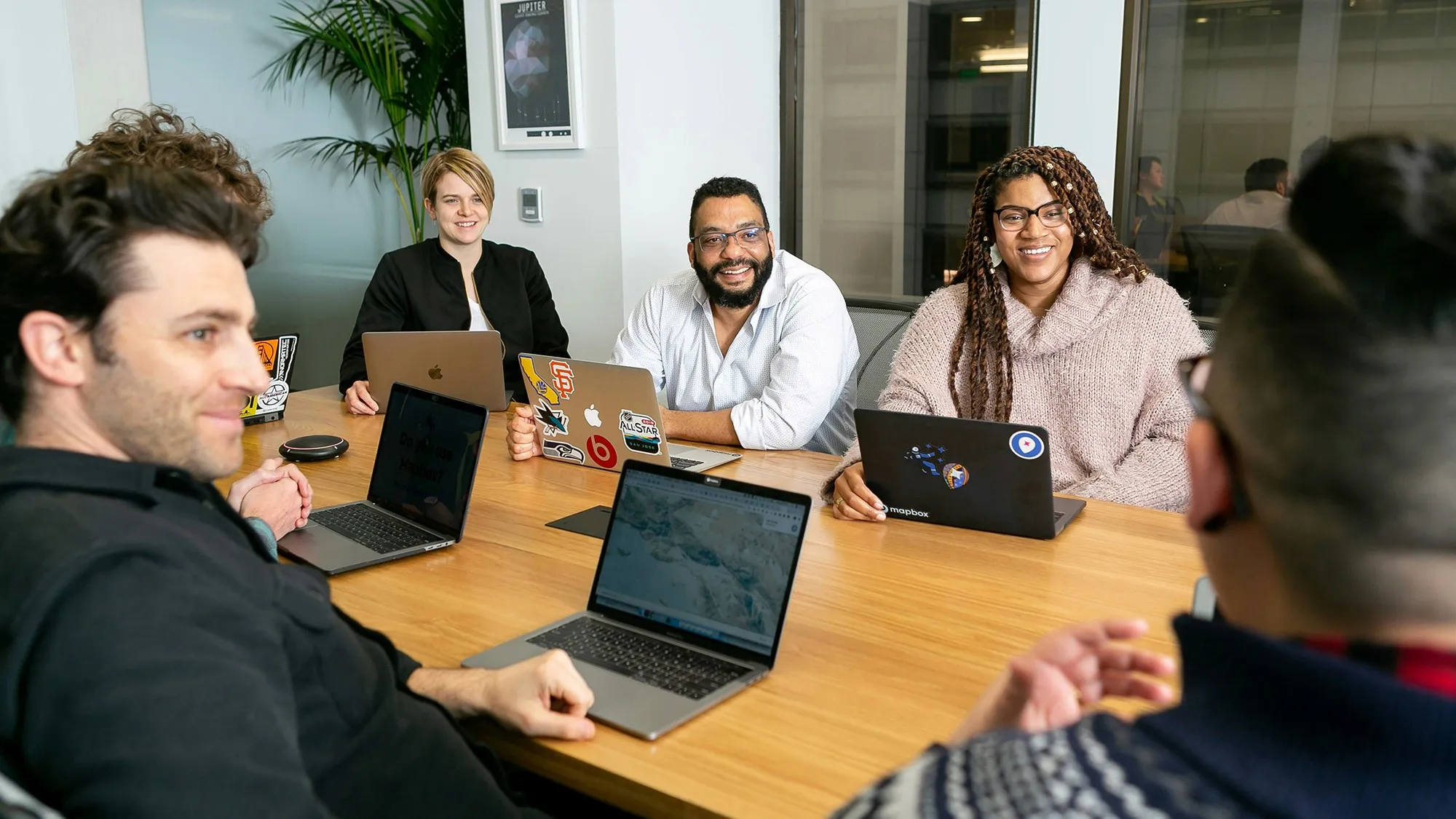A diverse group of working adults gathered around a conference table with laptops, smiling and engaged in discussion.