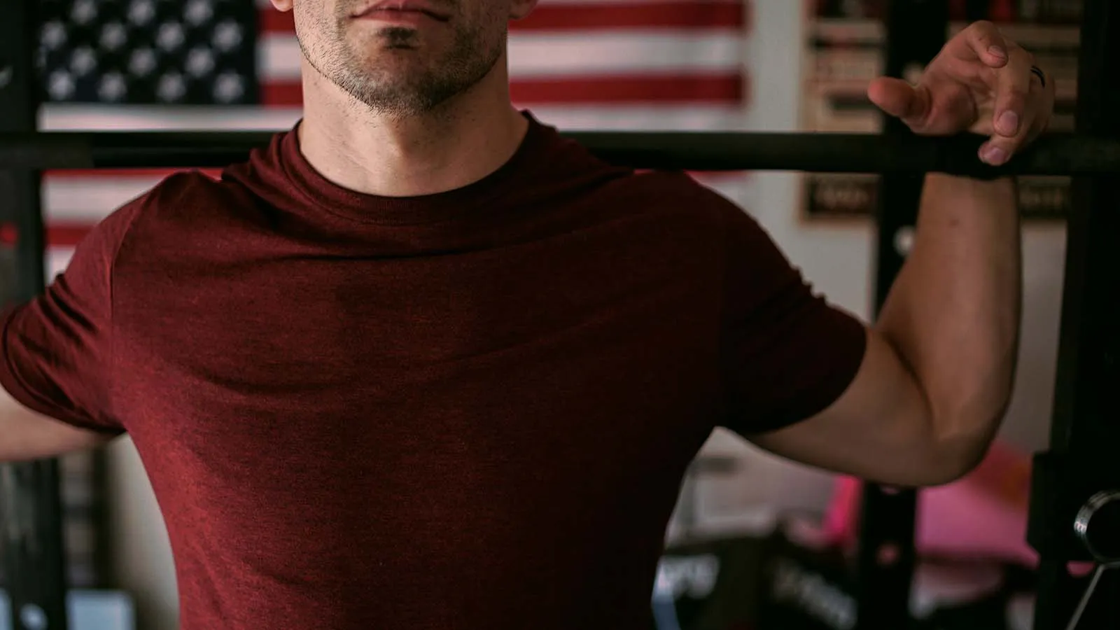 A man in a maroon t-shirt performing a barbell squat with an American flag in the background, representing how strength training supports healthy testosterone levels.