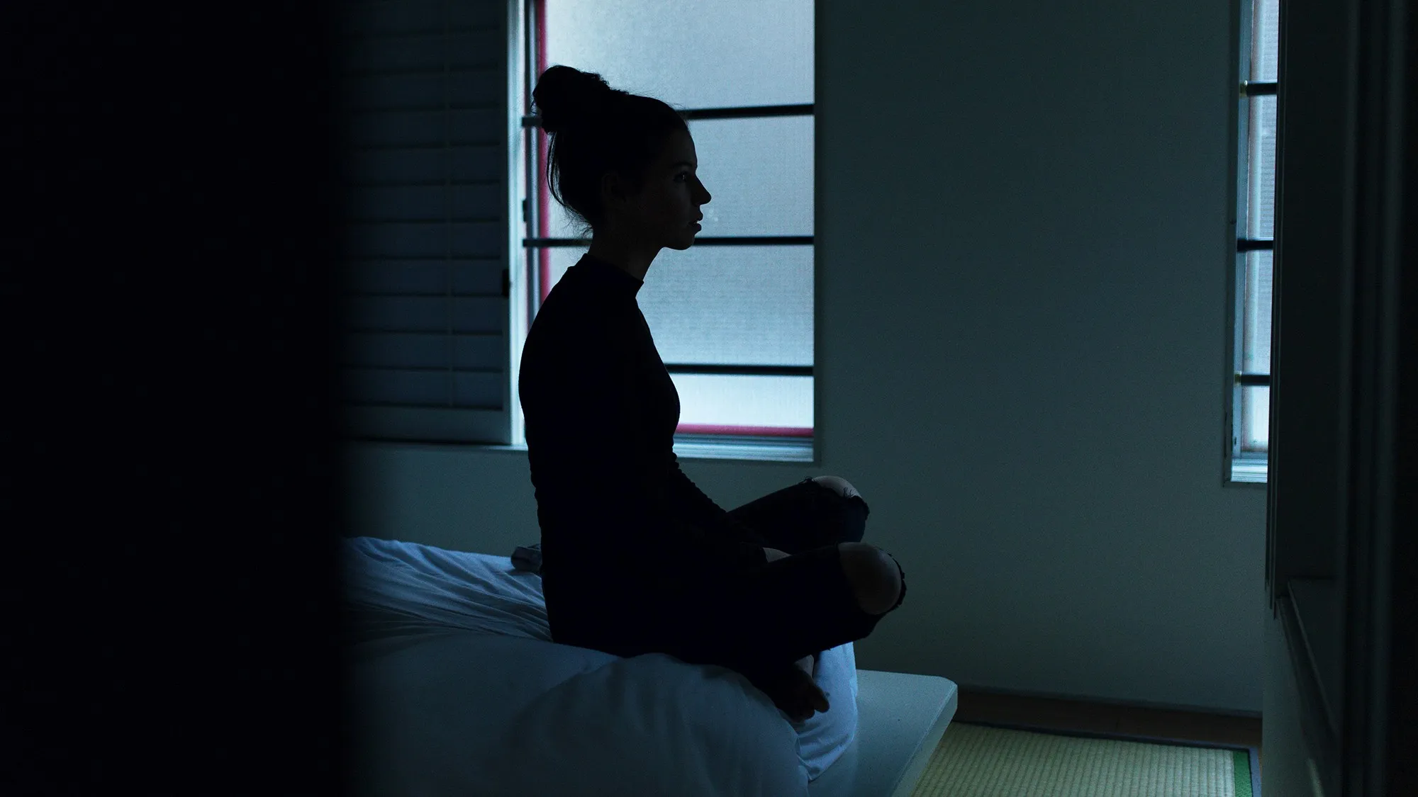 A silhouette of a woman sitting on her bed in a dark room, reflecting insomnia or a racing mind at bedtime.